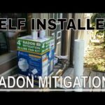 There are several items on a wooden deck including a box labeled as a radon mitigation in-line fan kit, multiple PVC pipes, and PVC fittings, suggesting preparation or the process of radon mitigation installation. The outdoor setting includes surrounding trees and a part of a house. Text overlaid on the image reads "SELF INSTALLED RADON MITIGATION."
