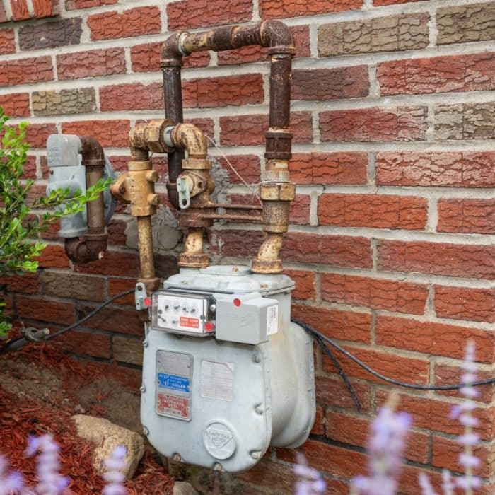 Gas meter on brick wall with rusted piping coming out the top too close to dirt