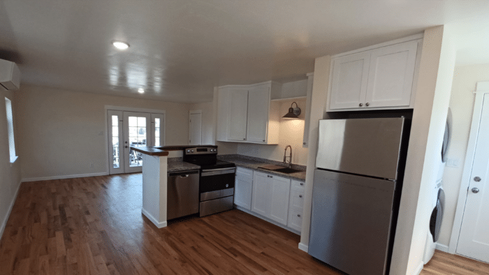 A kitchen with white cabinetry, stainless steel appliances, and hardwood flooring. A central island with a curved countertop is also visible, along with overhead and sconce lighting. Double doors leading outside are at the rear of the room.