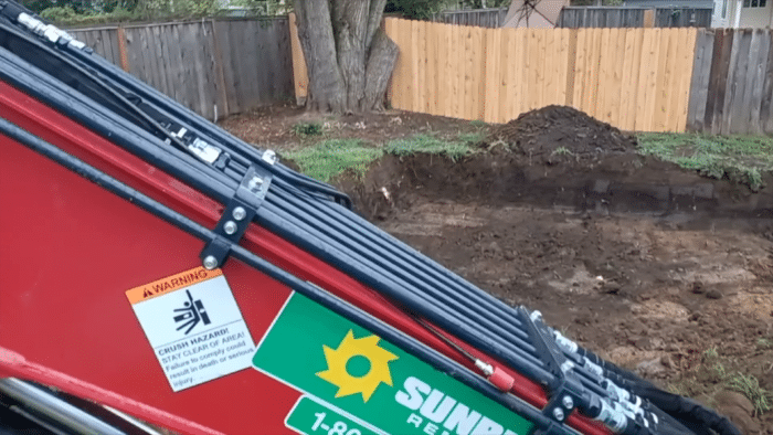 A partially visible red machinery with multiple cables and warning labels, positioned in front of a dug-up area of land with a newly installed wooden fence in the background.