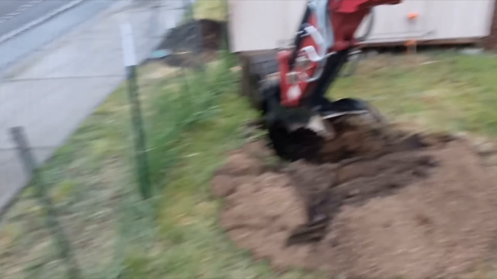 A red and black mini excavator is digging in a grassy area next to a concrete path. The soil is piled nearby and the scene is slightly blurred, indicating motion or low light conditions during the capture.